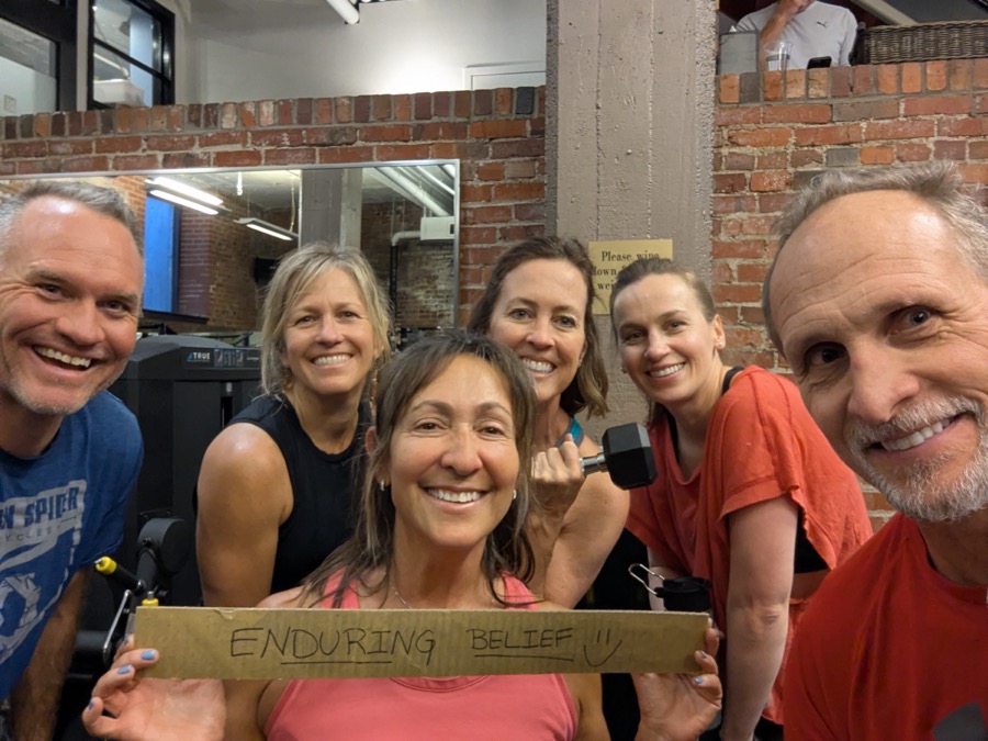 Alisa and five smiling clients after a session, holding a hand-lettered sign that reads Enduring Belief